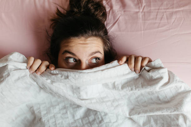 erschrockene und überraschte junge frau mit offenen augen versteckt gesicht unter decke, ziemlich erschrocken und neugierig mädchen gefühl schüchtern gucken aus bettdecke, abdeckung mit weißen blatt, kopf aus nächster nähe geschossen. ansicht von ob - sheet women bed sleeping stock-fotos und bilder