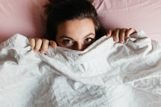 istock Playful young woman with beautiful eyes hiding face under blanket while lying in cozy bed, pretty curious girl feeling shy peeking from duvet, covering with white sheet, head shot close up. Top view 1177347360