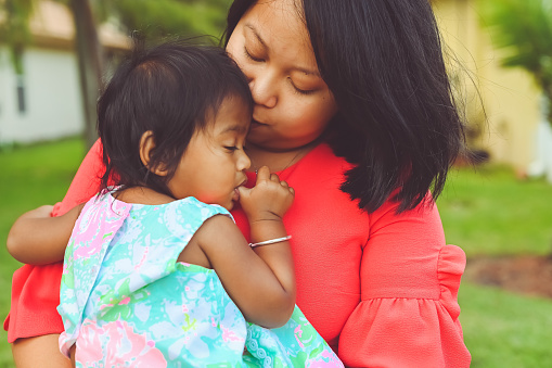 Picture of motherhood, sweet baby daughter sucks her thumb while her mother holds her close in a loving portrait. Mother is Vietnamese and daughter is Sri Lanken and Vietnamese. They are happy and heartwarming in a portrait of childhood and parenting