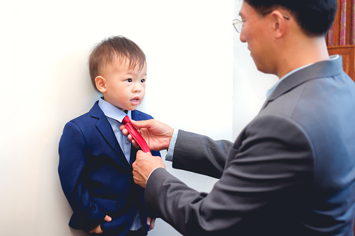 Asian father and son dressed in suits, Businessman helping his little child tying the necktie, Little kid looking and smiling at his dad, Happy Father's Day, Important role models concept