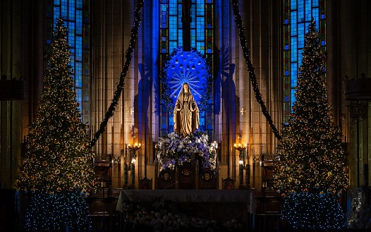 Saint Maria's sculpture - the Mother of God, in Catholic cathedral, in beams of blue and yellow color, on background of stained-glass window. Christmas decorations is visible.