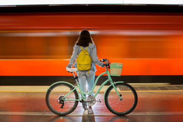 Teenager girl in jeans with bike standing on metro station Teenager girl in jeans with yellow backpack and bike standing on metro station, waiting for train, smiling, laughing. Orange train passing by behind the girl. Futuristic subway station. Finland, Espoo public transportation stock pictures, royalty-free photos & images