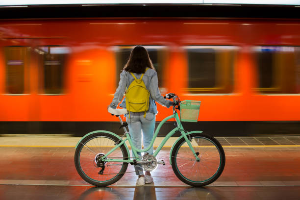 Teenager girl in jeans with bike standing on metro station Teenager girl in jeans with yellow backpack and bike standing on metro station, waiting for train, smiling, laughing. Orange train passing by behind the girl. Futuristic subway station. Finland, Espoo railroad station platform photos stock pictures, royalty-free photos & images