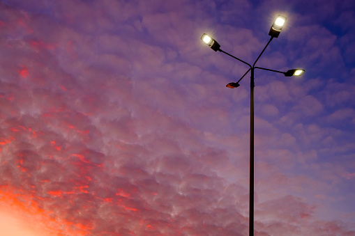 A modern street lamp shines against the backdrop of a colorful sunset sky. Many cirrus clouds are illuminated from below by the setting sun and cast in red.