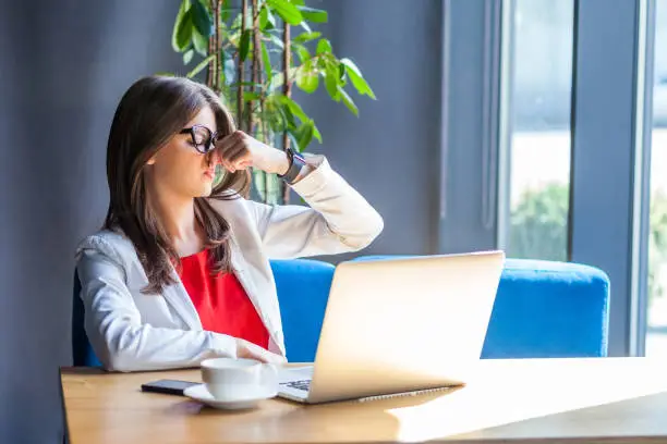 Bad smell. Portrait of dissatisfied beautiful stylish brunette young woman in glasses sitting pinching her nose and don't want to smelling. indoor studio shot, cafe, office background.