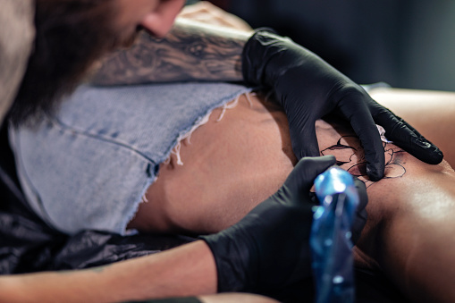 Young man tattooist doing tattoo on woman leg