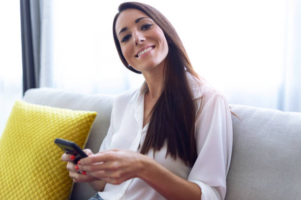 istock Pretty young woman looking at camera and using her mobile phone while sitting on sofa at home. 1177397617