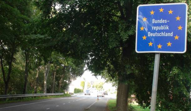 Roadside sign with EU stars marking entry into Germany, labeled "Bundesrepublik Deutschland," beside a tree-lined street.