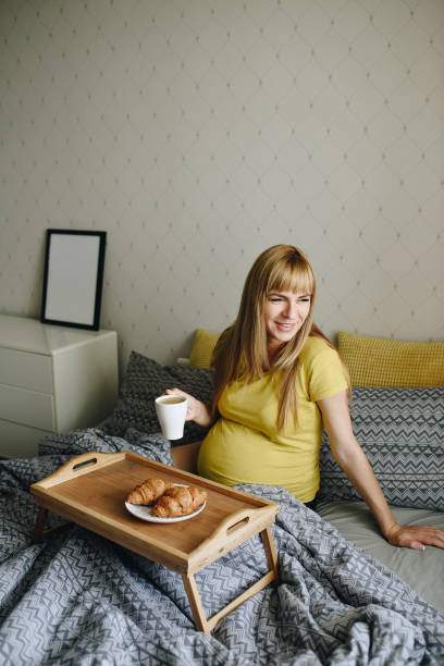 집에서 노란색 티셔츠에 임신 한 소녀 금발. 기적을 기다리고 있습니다. 임신. 사랑 희망. 회색 침구. 침실에 침대에 누워. 텍스트의 프레임입니다. 침대에서 아침 식사. 커피와 크루아상. 나무 � - human pregnancy miracle bed love 뉴스 사진 이미지