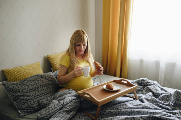 自宅で黄色のtシャツで妊娠中の女の子のブロンド。奇跡を待ってる妊娠。愛の希望灰色の寝具寝室のベッドに横たわっています。ベッドで朝食。クロワッサンとコーヒー。木製トレイ - human pregnancy miracle bed love ストックフォトと画像