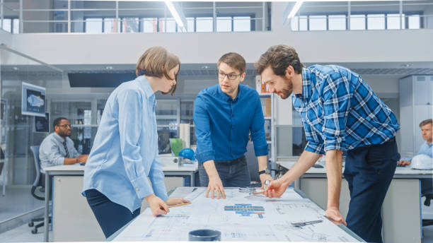 istock In the Industrial Engineering Facility: Female Designer Works with Industrial Engineer and Master Technician, They have Discussion, Analyse Engine Design Technical Drafts that are Lying on the Table 1177426350