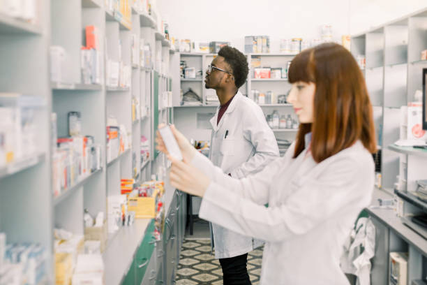 istock Two young multiethnical Pharmacists Working at modern pharmacy. Female Pharmacist Holding package with Pills, male pharmacist standing on the background. Pharmacists Wearing Medical Uniform. 1177434206