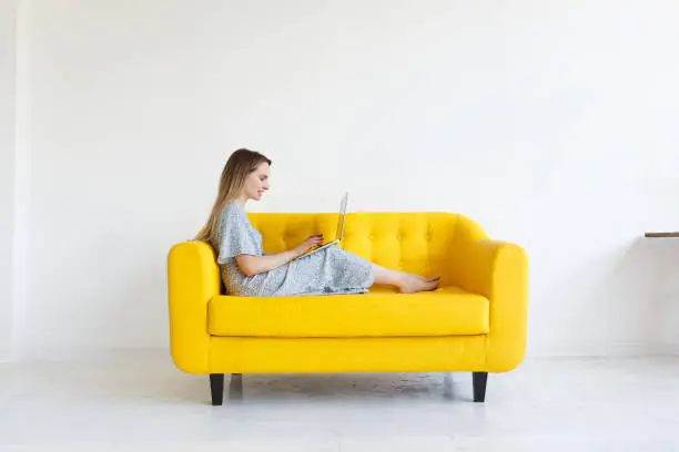 Photo of Side view. Beautifull charming girl checks email online, uses modern laptop computer, works remotely, uses wifi. Nice-looking woman smiling sits on yellow sofa at home over white background. Copyspace