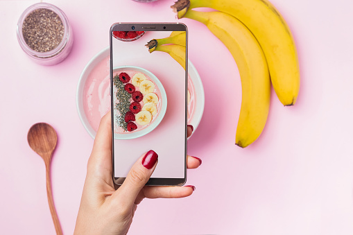 Woman's hand holding phone above the table with smoothie bowl and taking a picture of her breakfast.