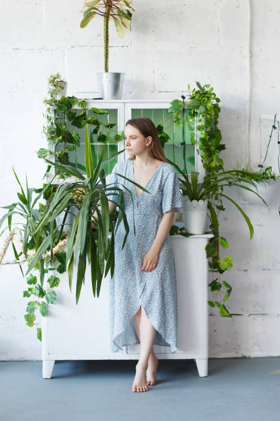 Young beautifull female model dressed blue casual dress posing surrounded by green plants and wondrous flowers in the botanical conservatory against white wall and sideboard. People, beauty and nature