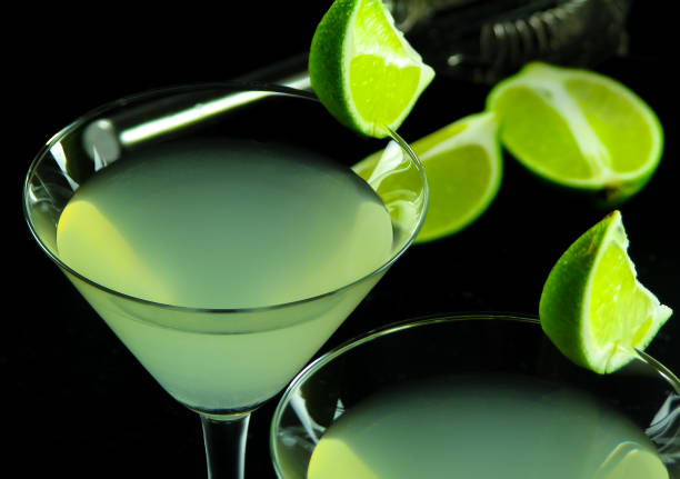 Alcoholic cocktail daiquiri light green color in cocktail glasses, decorated with lime slices close-up on the blurry background lime halves, strainer and dark surface