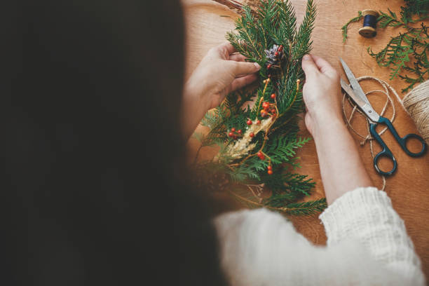istock Christmas wreath workshop. Hands holding fir branches, pine cones, berries, thread, scissors on wooden table. Making rustic christmas wreath. Authentic rural wreath 1177462523