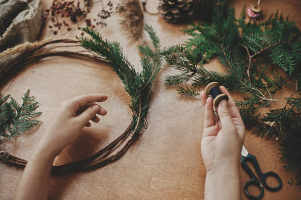 istock Hands holding thread and cedar branches , pine cones, scissors on wooden table. Details for workshop of making christmas wreath. Making rustic christmas wreath. 1177463131
