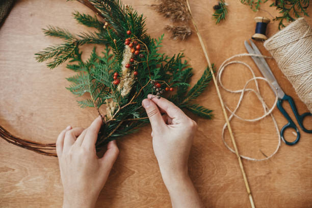 istock Christmas wreath workshop. Hands holding herbs, fir branches, pine cones, berries, thread, scissors on wooden table. Making rustic christmas wreath. Authentic rural wreath 1177464015