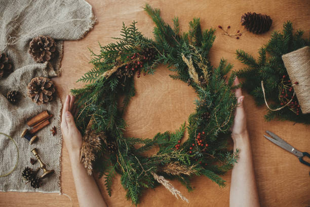 istock Rustic Christmas wreath, flat lay. Hands holding christmas wreath with fir branches, berries, pine cones, and thread, scissors on rural wooden table. Christmas wreath workshop 1177464262