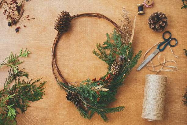 istock Rustic Christmas wreath, flat lay. Creative christmas wreath with fir branches, berries, pine cones and herbs with scissors and thread on rural wooden table. Happy holidays. Workshop 1177464500