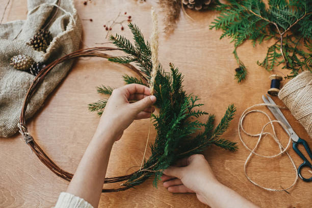 istock Hands holding herbs, fir branches, pine cones, berries, thread, scissors on wooden table. Making rustic christmas wreath. Christmas wreath workshop. Authentic rural wreath 1177464609