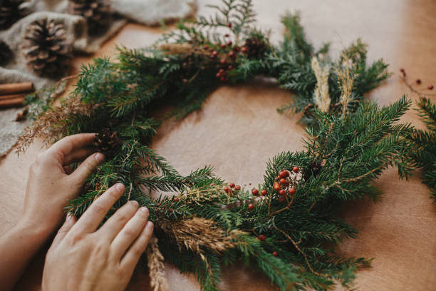 istock Hands holding christmas wreath with fir branches, berries, pine cones, and thread, scissors on rural wooden table. Christmas wreath workshop. Rustic Christmas wreath. 1177464754