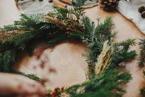 istock Christmas wreath workshop. Hands holding berries and  fir branches, pine cones, thread on wooden table. Authentic stylish still life. Making rustic Christmas wreath. 1177465408