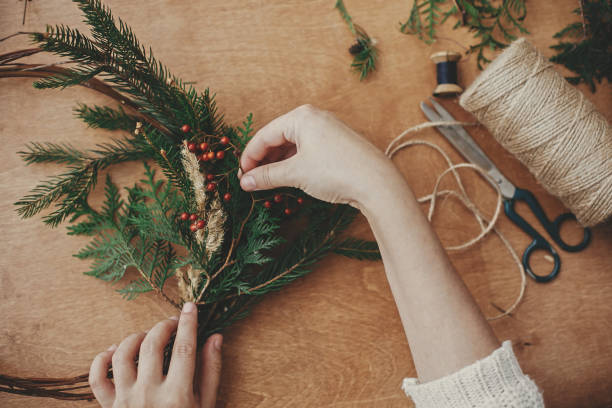 istock Christmas wreath workshop. Hands holding fir branches, pine cones, berries, thread, scissors on wooden table, flat lay. Making rustic christmas wreath. Authentic rural wreath 1177466295