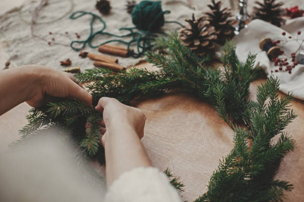 istock Making rustic Christmas wreath. Hands holding fir branches, and pine cones, thread, berries, scissors on wooden table. Christmas wreath workshop. Authentic stylish still life 1177466321