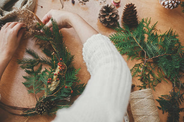 istock Christmas wreath workshop. Hands holding fir branches, pine cones, berries, thread, scissors on wooden table. Making rustic christmas wreath. Authentic rural wreath 1177467439
