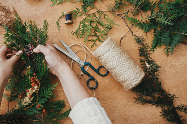 istock Christmas wreath workshop. Hands holding fir branches, pine cones, berries, thread, scissors on wooden table, flat lay. Making rustic christmas wreath. Authentic rural wreath 1177467660