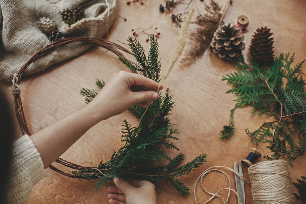istock Christmas wreath workshop. Hands holding herbs, fir branches, pine cones, berries, thread, scissors on wooden table. Making rustic christmas wreath. Authentic rural wreath 1177467813