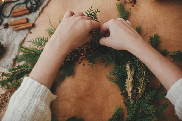 istock Hands holding fir branches, and pine cones, thread, berries, scissors on wooden table. Making rustic Christmas wreath flat lay. Christmas wreath workshop. Authentic stylish still life 1177468399