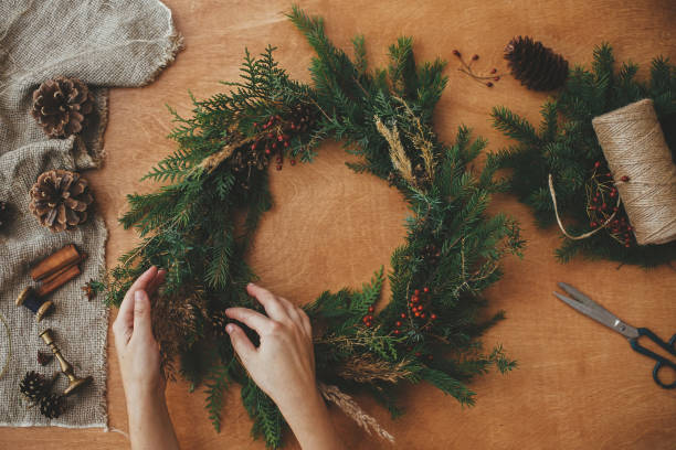 istock Hands holding christmas wreath with fir branches, berries, pine cones, and thread, scissors on rural wooden table. Rustic Christmas wreath, flat lay. Christmas wreath workshop 1177468951