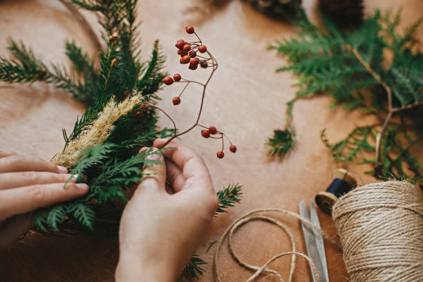 istock Christmas wreath workshop. Hands holding berries, fir branches, pine cones, thread, scissors on wooden table. Making rustic christmas wreath. Authentic rural wreath 1177468987