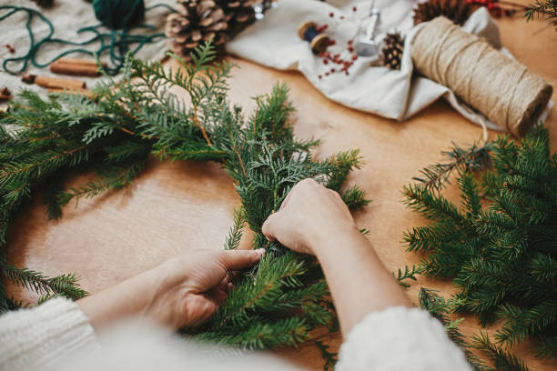 istock Making rustic Christmas wreath. Hands holding fir branches, and pine cones, thread, berries, scissors on wooden table. Christmas wreath workshop. Authentic stylish still life 1177469100