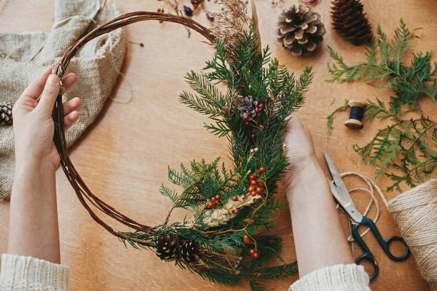 istock Hands holding rustic christmas wreath with pine cones, berries, fir branches, thread, scissors on wooden table.  Authentic rural wreath. Christmas wreath workshop. 1177469115