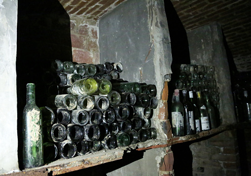 Very ancient wine bottles with dusty shelves stand in an abandoned dark cellar of a French winery building.