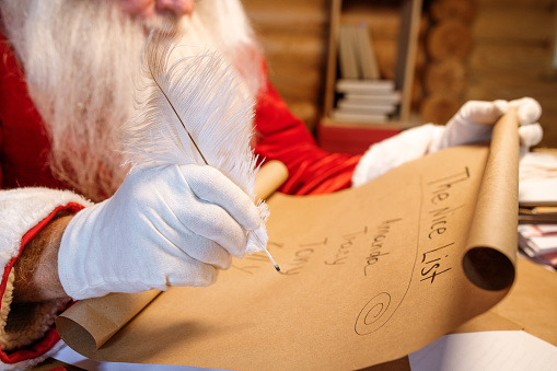 Gloved hand of Santa holding white plume over nice list on unrolled paper while getting ready for Christmas