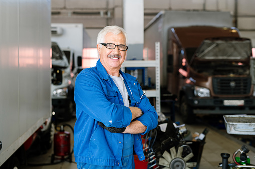 Happy mature cross-armed technician of car maintenance service looking at you on background of workshop interior