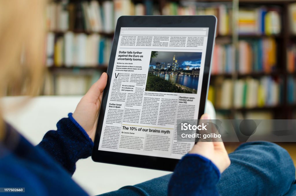 Young woman reading the news on a modern tablet computer, while sitting in her living room A digital newspaper on a modern tablet computer held by a young woman, who is reading the day’s news. The photo was taken by looking over the model's shoulder. A bookshelf is visible in the background of the picture. The newspaper on the device screen was created by myself in Indesign,  I own the copy right of the design and layout. The image of Frankfurt on the device is my own, I own the copy right. The text is all lorem ipsum except for the titles, which are generic and entirely fictional – these were written by myself. Newspaper Stock Photo