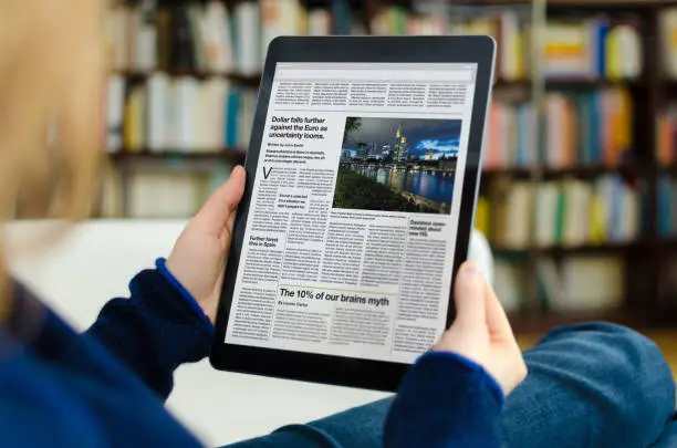A digital newspaper on a modern tablet computer held by a young woman, who is reading the day’s news. The photo was taken by looking over the model's shoulder. A bookshelf is visible in the background of the picture. The newspaper on the device screen was created by myself in Indesign,  I own the copy right of the design and layout. The image of Frankfurt on the device is my own, I own the copy right. The text is all lorem ipsum except for the titles, which are generic and entirely fictional – these were written by myself.