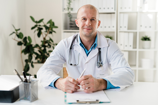 Young successful clinician in eyeglasses and whitecoat sitting by desk in medical office in front of camera
