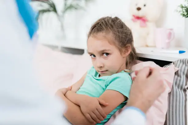 Reluctant sick little girl looking at spoon with medicine held by doctor while sitting in bed at home