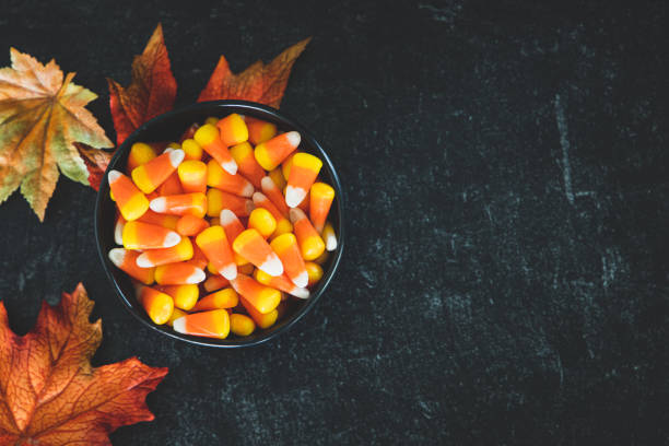 A Small bowl of candy corn on a table with some autumn leaves A Small bowl of candy corn on a table with some autumn leaves as decoration. Perfect for Halloween and autumn fall decoration. Black onyx background. candy corn stock pictures, royalty-free photos & images