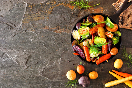 Cast iron skillet of roasted autumn vegetables, above view side border over a dark stone background