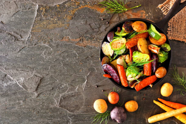 Cast iron skillet of roasted autumn vegetables, above view side border on a dark slate background Cast iron skillet of roasted autumn vegetables, above view side border over a dark stone background cooking pan overhead stock pictures, royalty-free photos & images