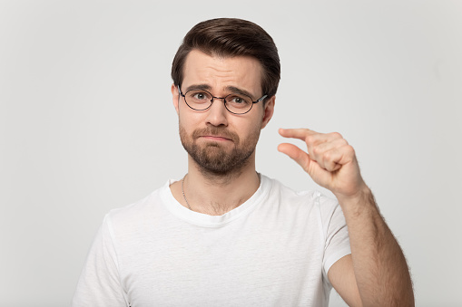 Disappointed millennial man in glasses showing small size length with a pity, isolated on grey white studio background. Head shot portrait young guy looking at camera showing measurement with fingers.