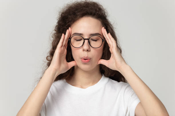 Tired woman in eyewear massaging temples, trying to reduce stress. Head shot close up portrait young tired woman in eyewear massaging temples, trying to reduce stress and calm down, breathing fresh air, isolated on grey white studio background, self-control concept. exhaling stock pictures, royalty-free photos & images